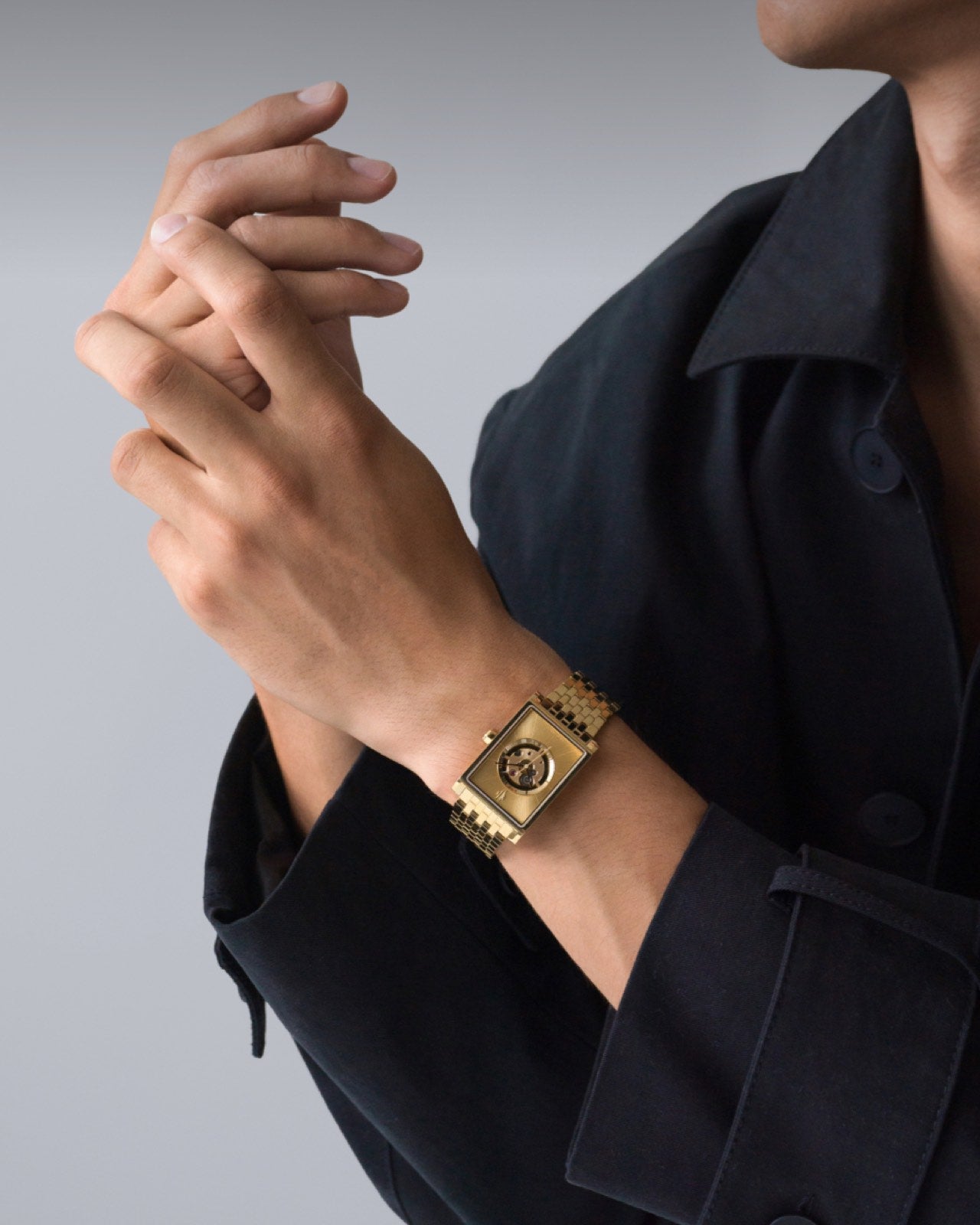 Gold rectangular automatic watch worn on man's wrist with black jacket